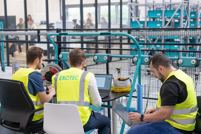 A group of men wearing Exotec vests working on laptops inside a warehouse.