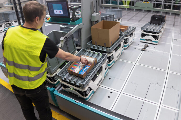 E-Commerce Fulfillment: Ein Kommissionierer greift in einen Karton der auf einem Tablar auf einem Lagerroboter steht