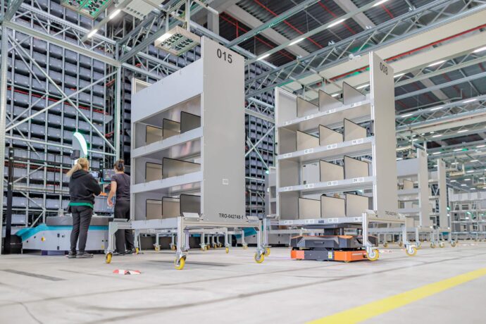 Zwei Mitarbeiterinnen an einer Packstation im Lager Dosenbach-Ochsner in Luterbach, im Vordergrund Safelog-AGVs, die mobile Kommissionierregale autonom durch die Halle transportieren, im Hintergrund das Hochregallager des Skypod®-Systems.