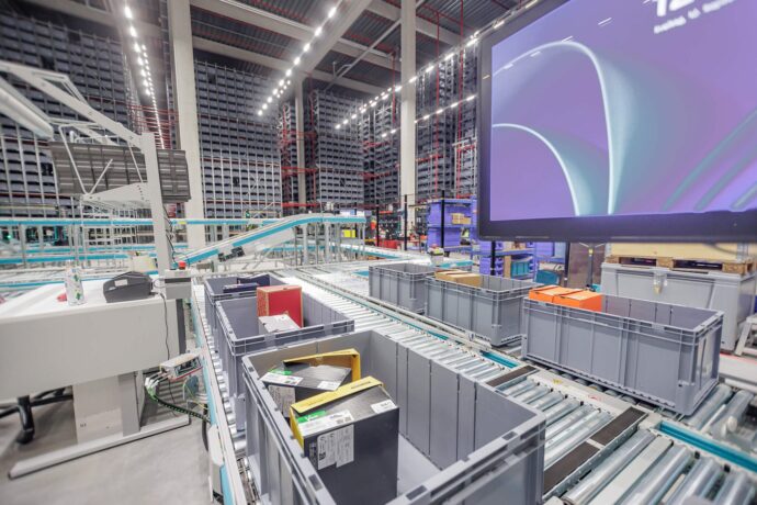 Rollenförderbänder mit grauen Transportbehältern und Schuhkartons, im Hintergrund das bis zur Decke reichende Hochregallager des Skypod®-Systems von Exotec